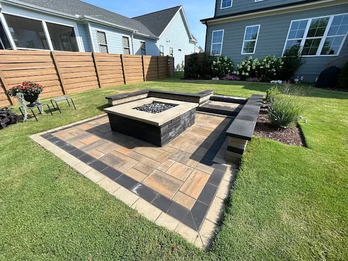 Rectangular paver patio with a black square fire pit at its center, capped seat walls on two sides and a contrasting stone border, set into a manicured lawn behind a blue-sided home.