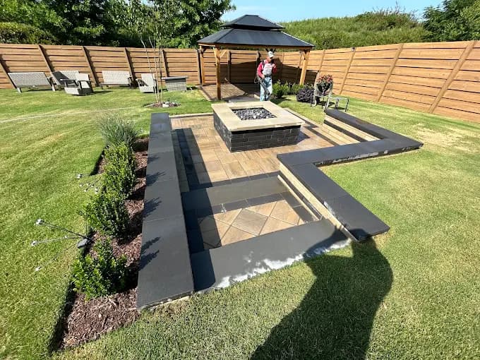 Backyard paver patio with a square fire pit and low seat walls, joined by a dark paver walkway across the lawn to a pergola-covered seating area inside a horizontal wood privacy fence.