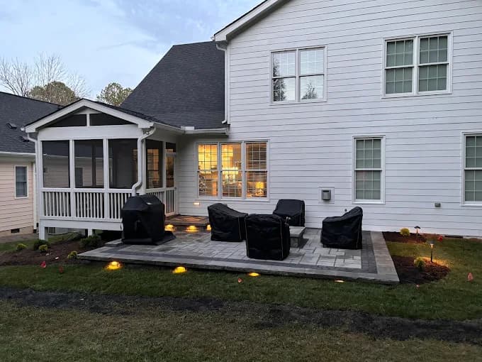 Paver patio at dusk beside a screened porch, with covered outdoor furniture and warm low-voltage landscape lights washing the edge of the planting bed.