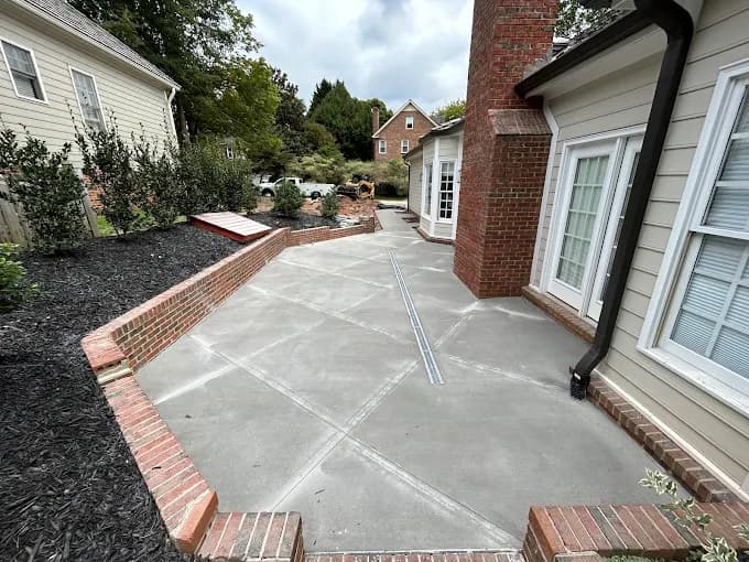 Broom-finish concrete patio with a soldier-course brick border wrapping the back of a brick home, with a linear trench drain set flush into the slab to carry water off the surface.