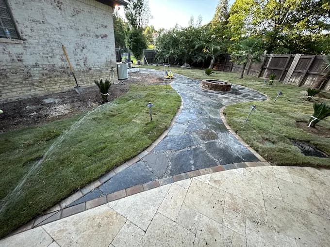 Wide flagstone walkway curving through a freshly planted backyard toward a round stone fire pit, with new mulch beds and young shrubs on both sides.