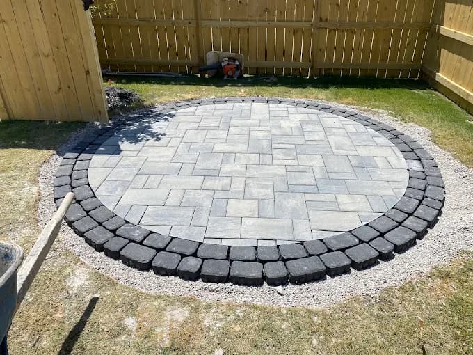Circular paver patio under construction, a light gray paver field ringed by a charcoal cobblestone border, laid over a compacted gravel base beside a wood fence.