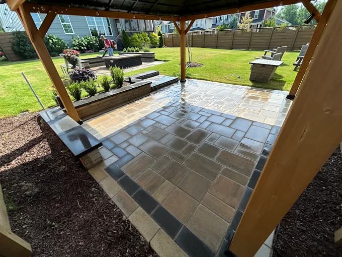 Flagstone patio under a heavy cedar pergola, edged by a low seat wall and fresh mulch beds, looking out across the lawn to Adirondack chairs on the far side.