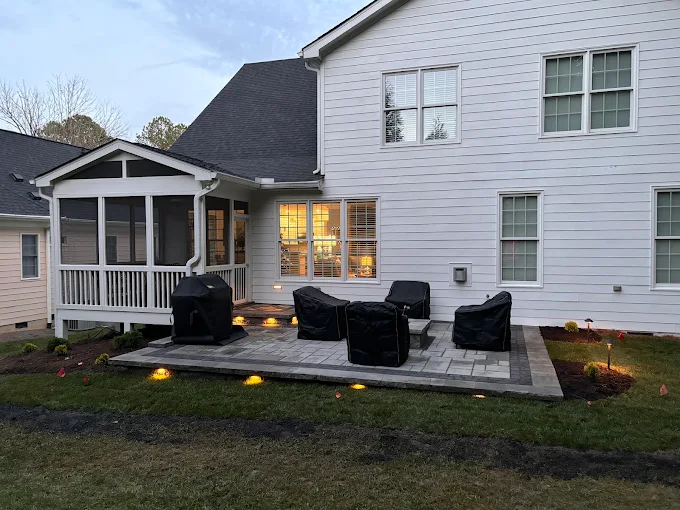 Paver patio at dusk beside a screened porch, with covered outdoor furniture and warm low-voltage landscape lights washing the edge of the planting bed.