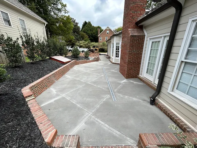 Broom-finish concrete patio with a soldier-course brick border wrapping the back of a brick home, with a linear trench drain set flush into the slab to carry water off the surface.