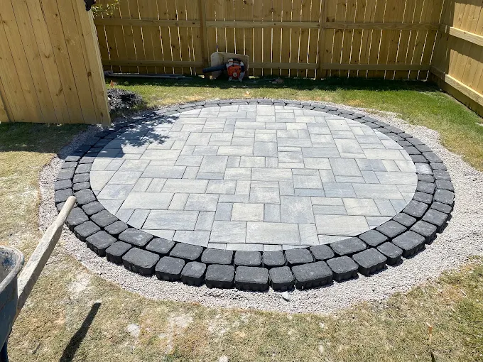 Circular paver patio under construction, a light gray paver field ringed by a charcoal cobblestone border, laid over a compacted gravel base beside a wood fence.