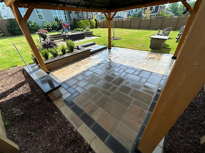 Flagstone patio under a heavy cedar pergola, edged by a low seat wall and fresh mulch beds, looking out across the lawn to Adirondack chairs on the far side.